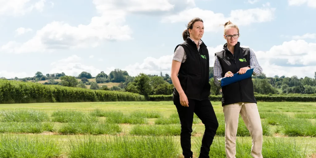 Dr Joanna Matthews and Grace Welling are standing among grass and forage trial plots at Germinal Horizon Wiltshire on a sunny day with blue sky and white clouds.
