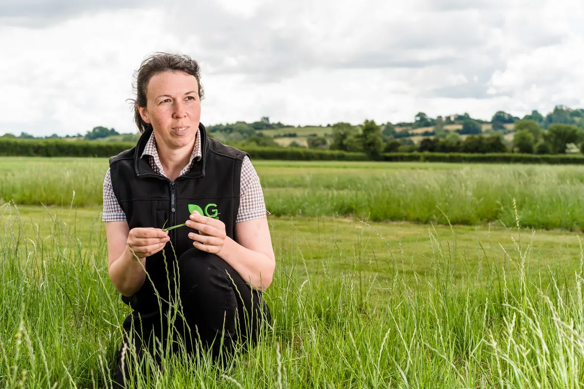Dr Joanna Matthews is crouching in a grass and forage trial plots at Germinal Horizon Wiltshire on a sunny day with blue sky and white clouds.
